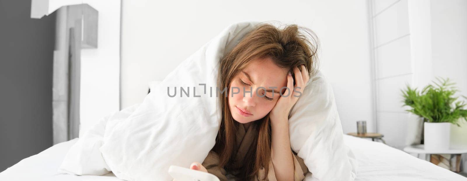 Depressed woman in bed, lying under blanket and looking at phone with sad, complicated face.