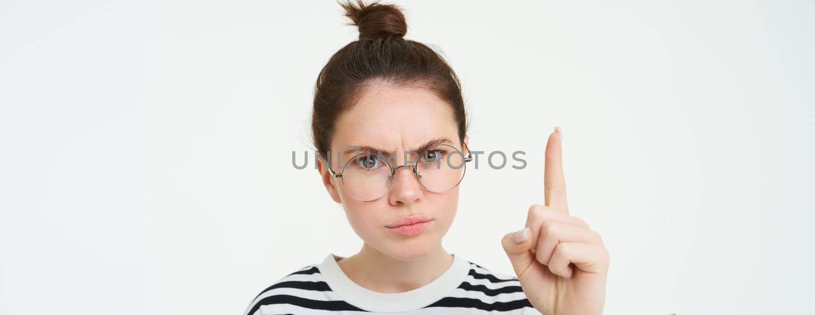 Portrait of young woman, tutor in glasses, pointing finger up and frowning, showing smth important, standing over white background.