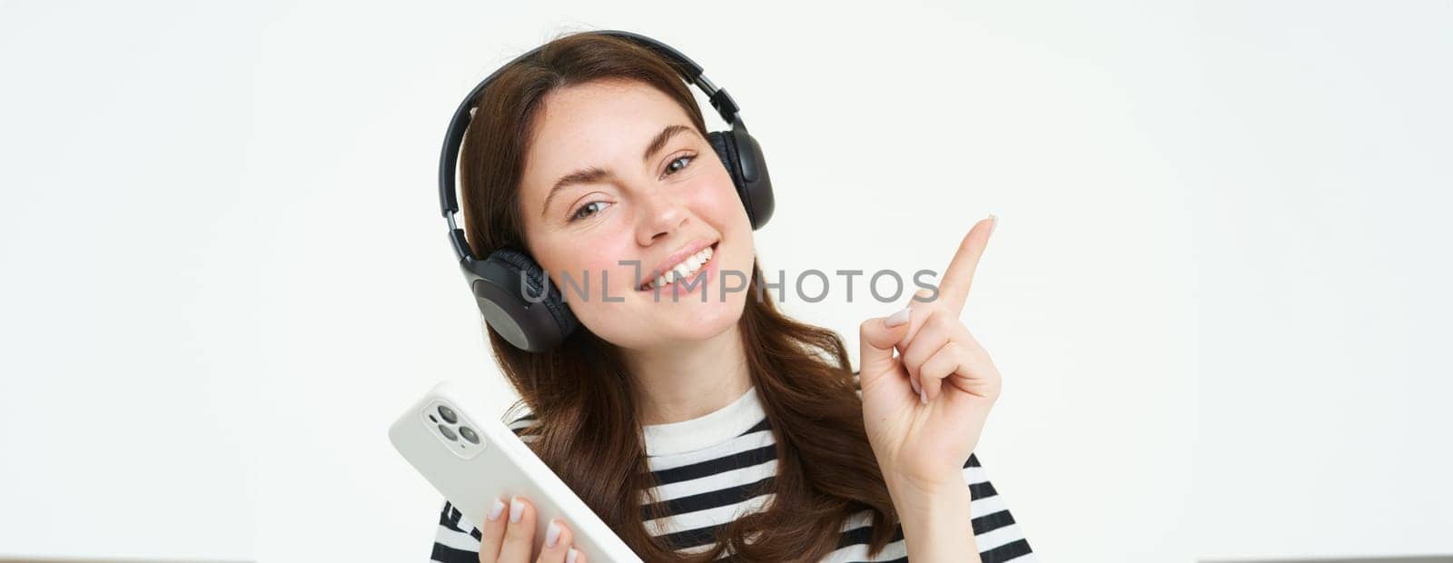 Portrait of smiling student, girl in headphones, holding mobile phone, pointing left, showing advertisement, store offer, white background by Benzoix