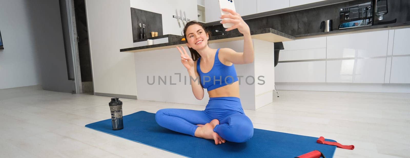 Young smiling woman, fitess girl doing workout from home, sits on yoga mat with smartphone, taking selfie, records sport vlog indoors.
