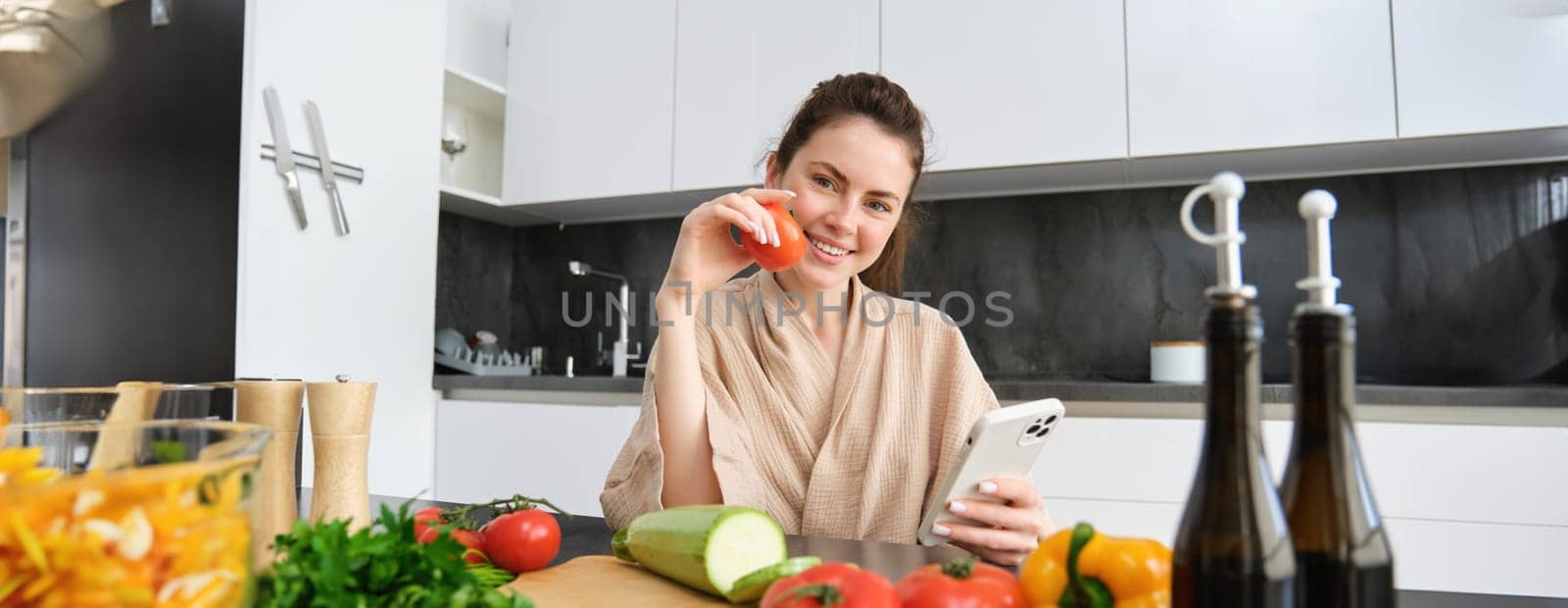 Portrait of young woman ordering groceries on smartphone app, holding tomato, sitting near chopping board with vegetables. Girl looking for recipe online, using mobile phone.