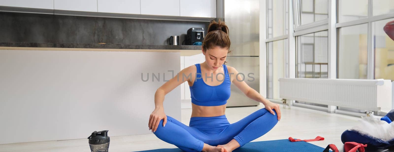Girl focused on her training session, workout from home, stretches her legs in yoga lotus pose, sits on fitness mat in sportsbra and leggings by Benzoix
