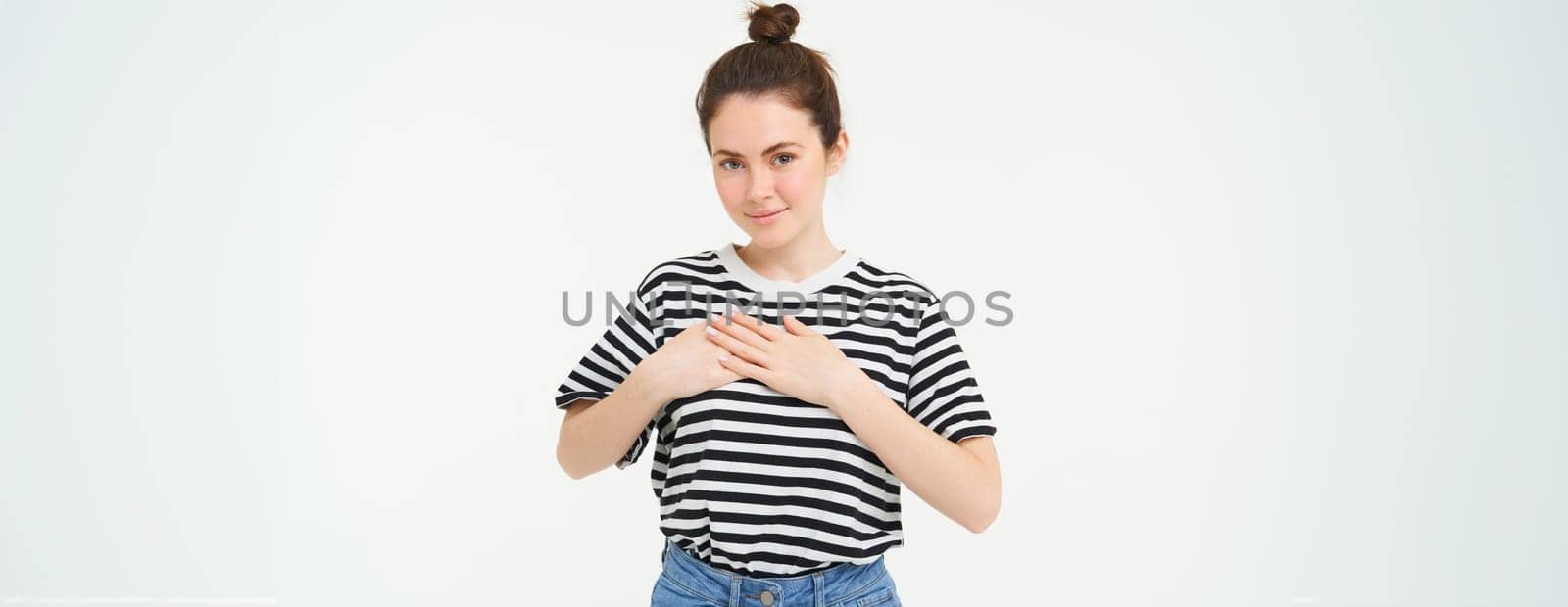 Young smiling woman, feels gratitude, thankful for something, holds hands on heart and looks tender at camera, standing over white background.