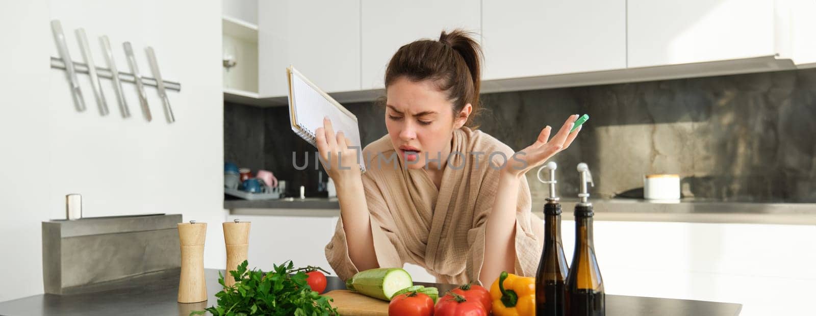 Portrait of woman cant cook, looking confused while making meal, holding recipe book, checking grocery list and staring frustrated at camera, standing near vegetables in the kitchen by Benzoix