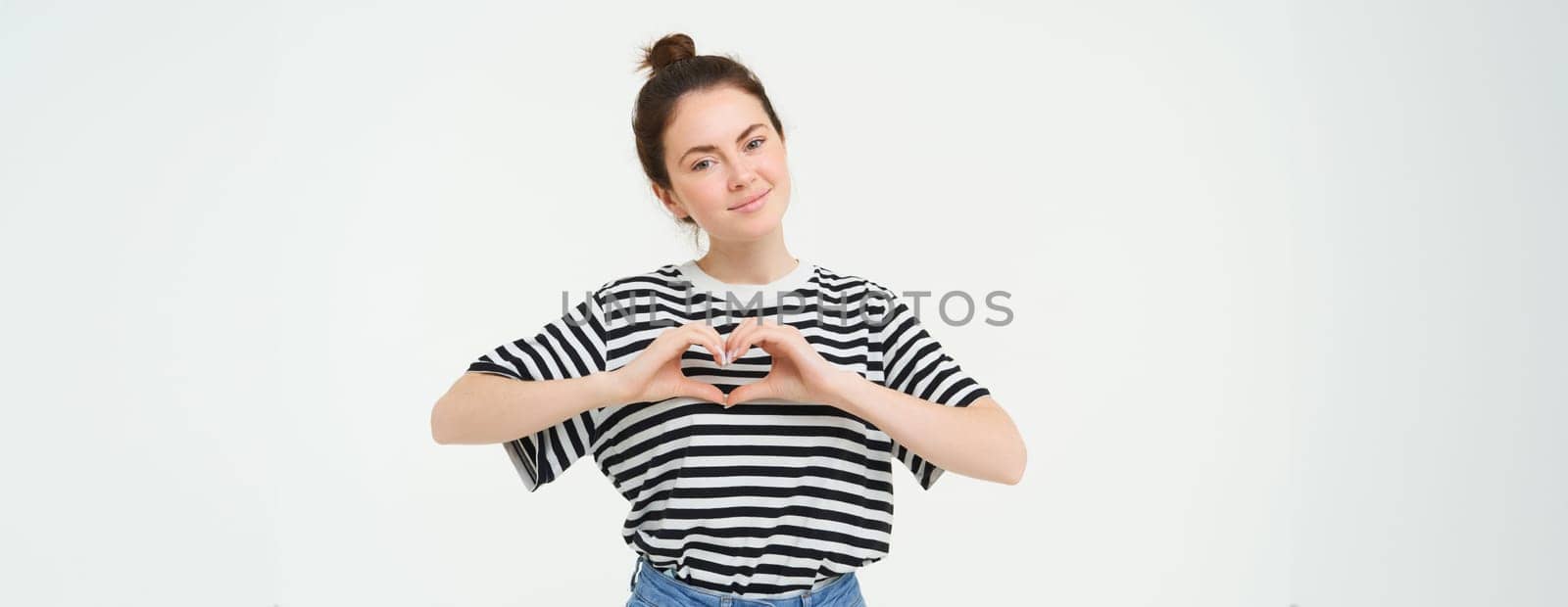 Portrait of beautiful girl shows heart sign, love gesture, like something, stands over white background. Copy space