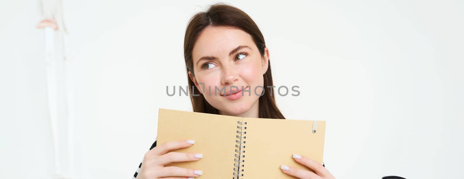 Portrait of beautiful young woman, holding daily planner, writing in her diary, making notes in notebook and smiling, dreaming about something, standing thoughtful against white background.