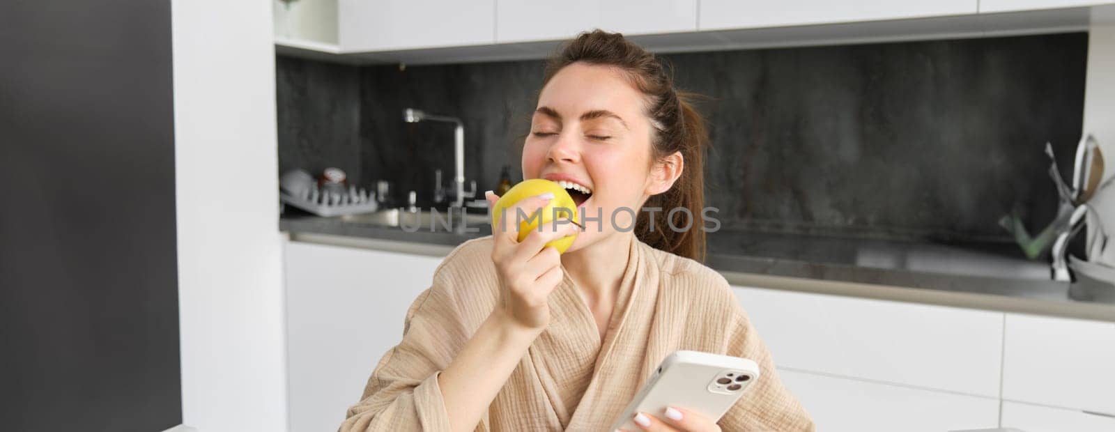 Portrait of happy, beautiful woman smiling, eating an apple in the kitchen, sitting at home in bathrobe, holding smartphone, using mobile phone.