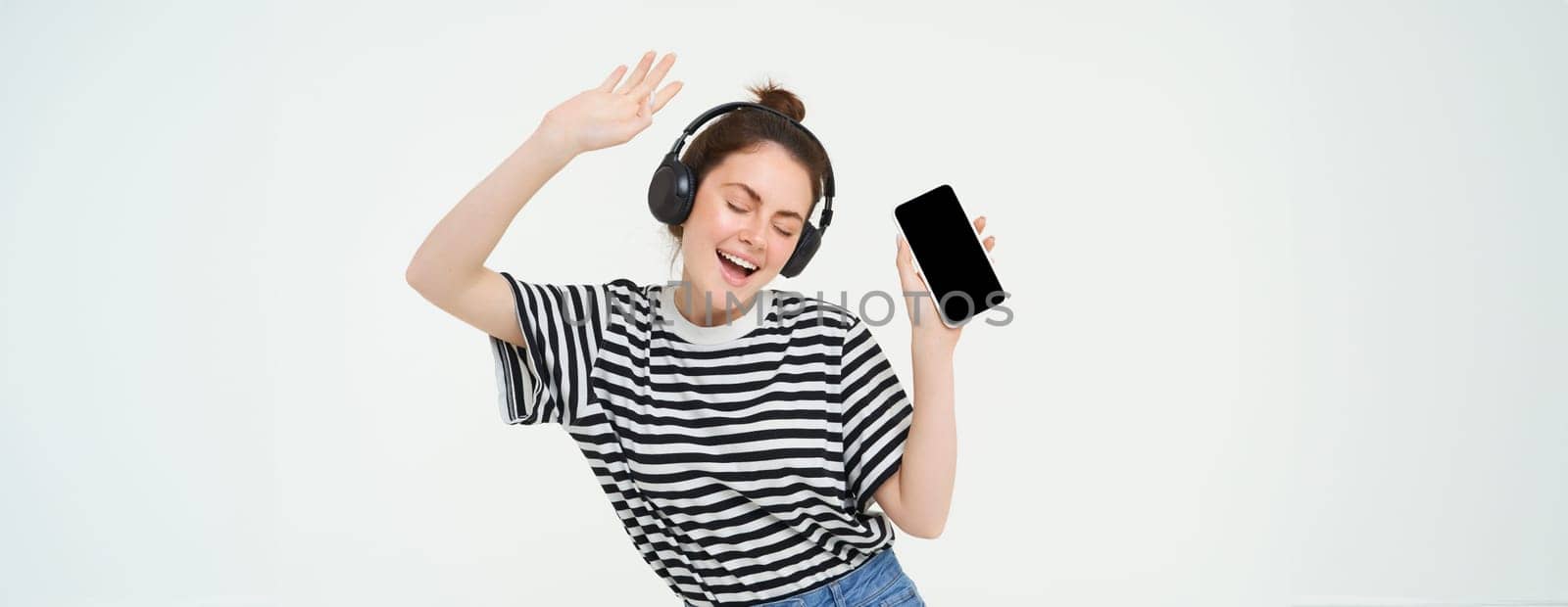 Portrait of stylish young woman dancing in wireless earphones, using headphones and mobile phone music app to listen to favourite songs, posing over white background.