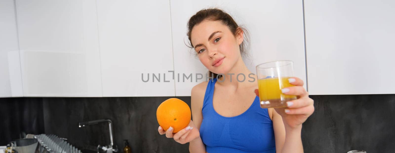 Close up portrait of sportswoman, fitness girl holding glass of fresh juice and an orange in hands, smiling at camera, standing in kitchen. Workout and healthy lifestyle concept