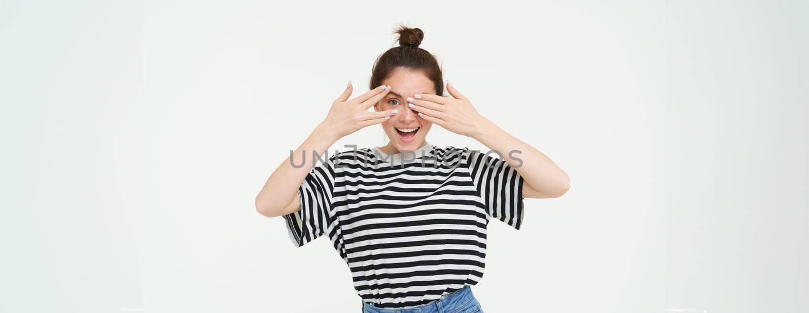 Happy young woman, bday girl peeking through fingers, holds hands on eyes, waiting for surprise, stands over white background by Benzoix