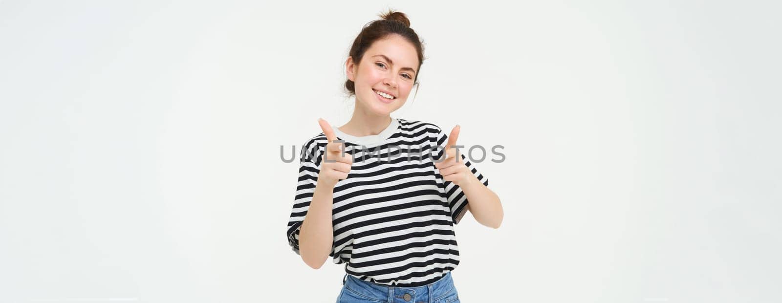 Portrait of cute girl, student shows thumbs up, recommends product, approves choice, likes item, stands over white background.