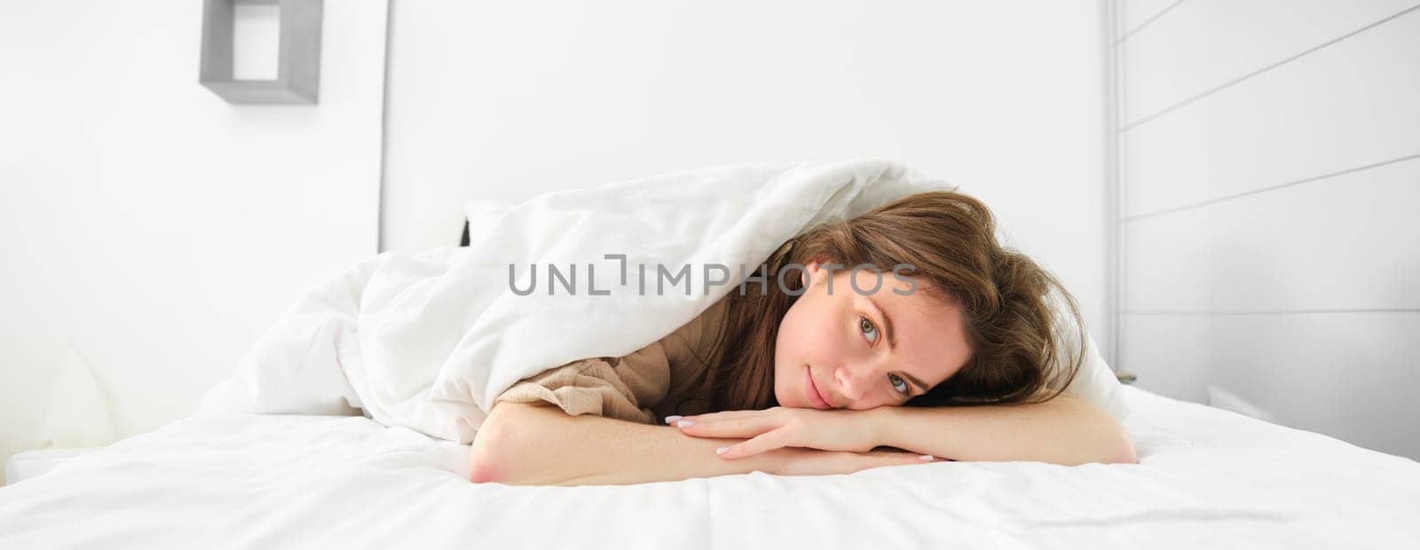 Beautiful young woman, lying in her bed under blanket, looking at camera with romantic, calm smiling face.