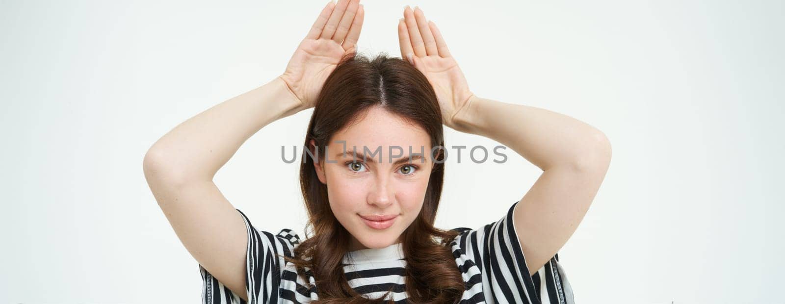 Image of cute brunette girl shows animal eats with hands on top of head, smiling and looking happy, white background.
