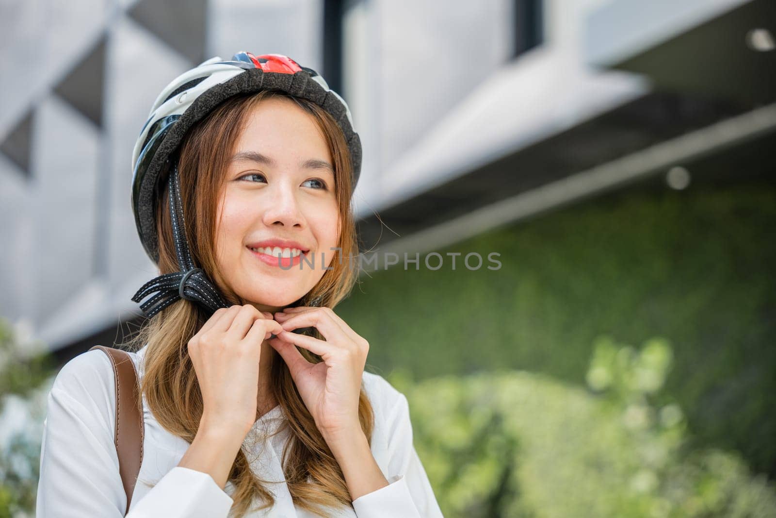 Close up smiling woman wearing helmet exercise outdoors with bicycle on street, Asian young businesswoman putting biking helmet for safety prepared cyclists around building go to work, Bike commuting