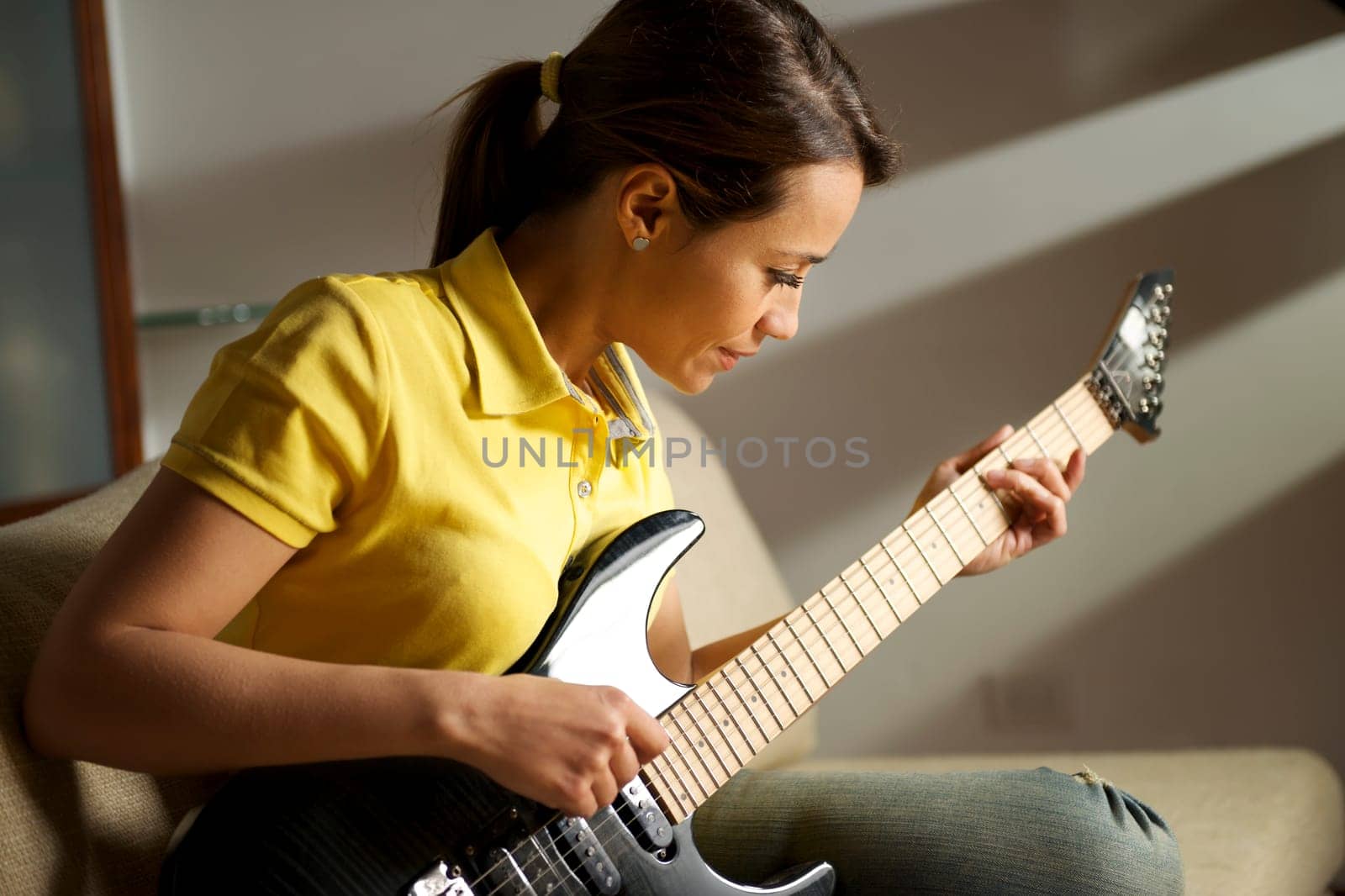 beautiful mid adult woman playing electric guitar at home, sitting on sofa