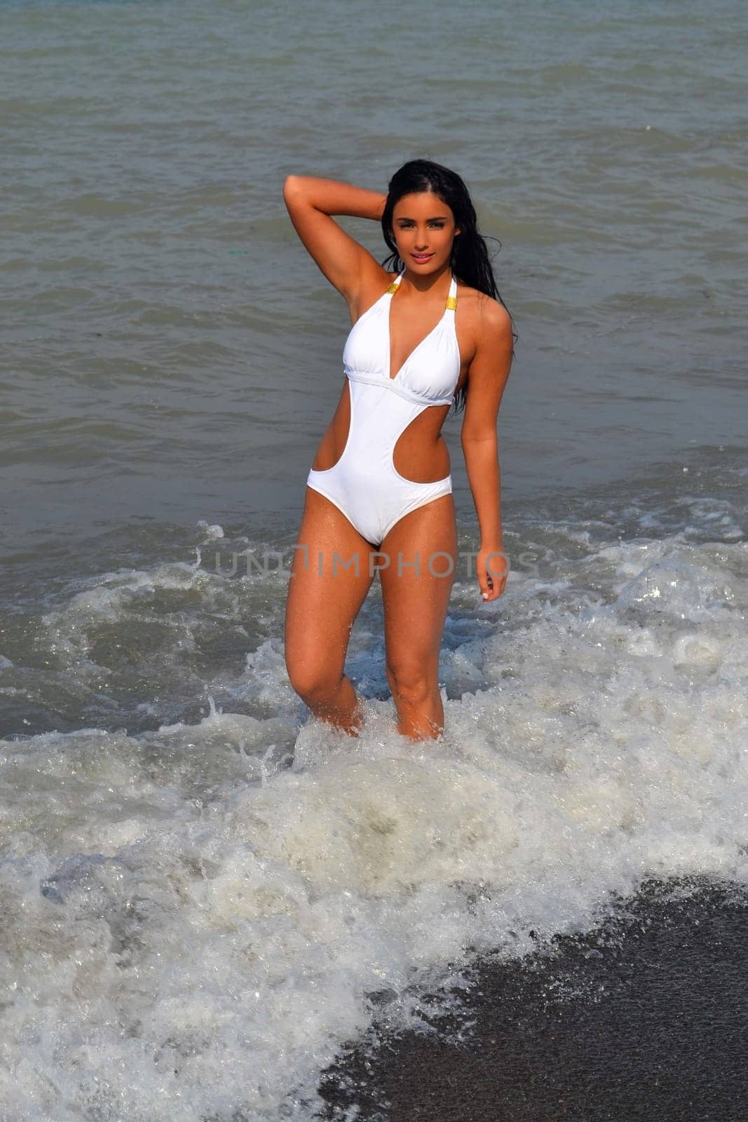 A gorgeous young woman in a white bikini and long black hair standing.in a wave on the shore of lake Ontario, having a good time..