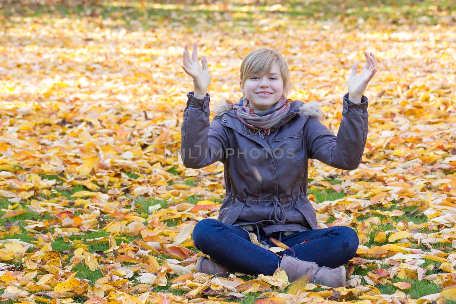 smiling girl sitting on fallen leaves in autumn park and rising up hands