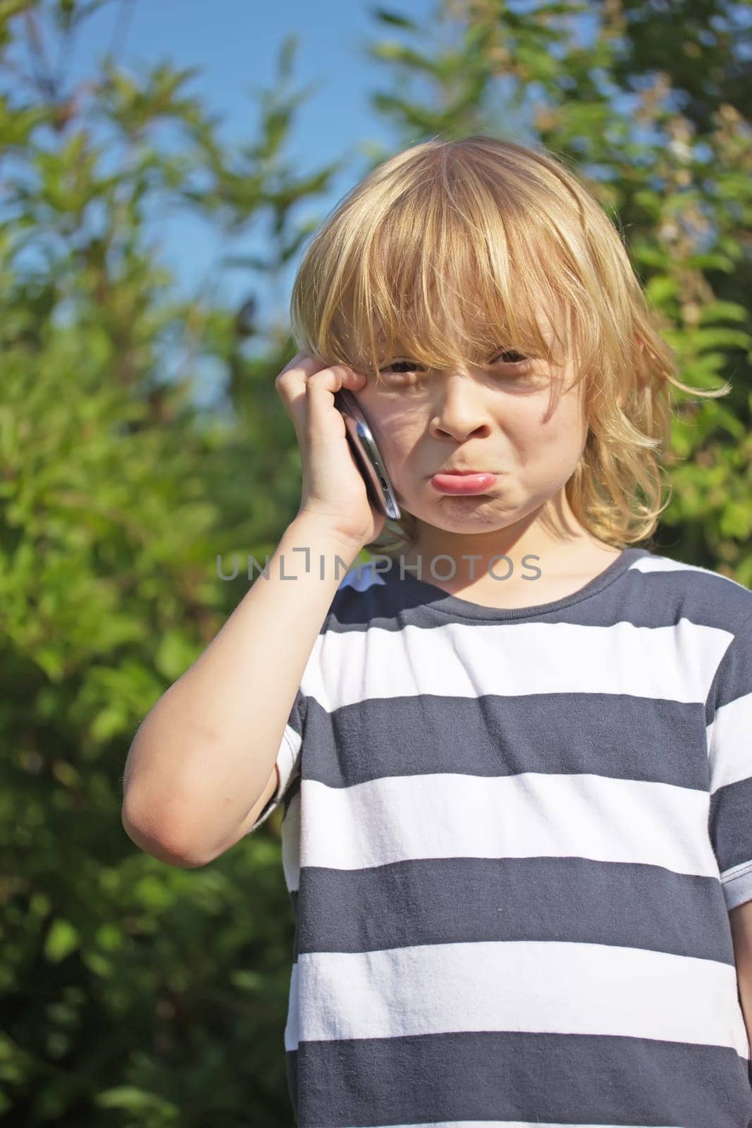 Annoyed blond boy in a stripped shirt  is calling outdoors.