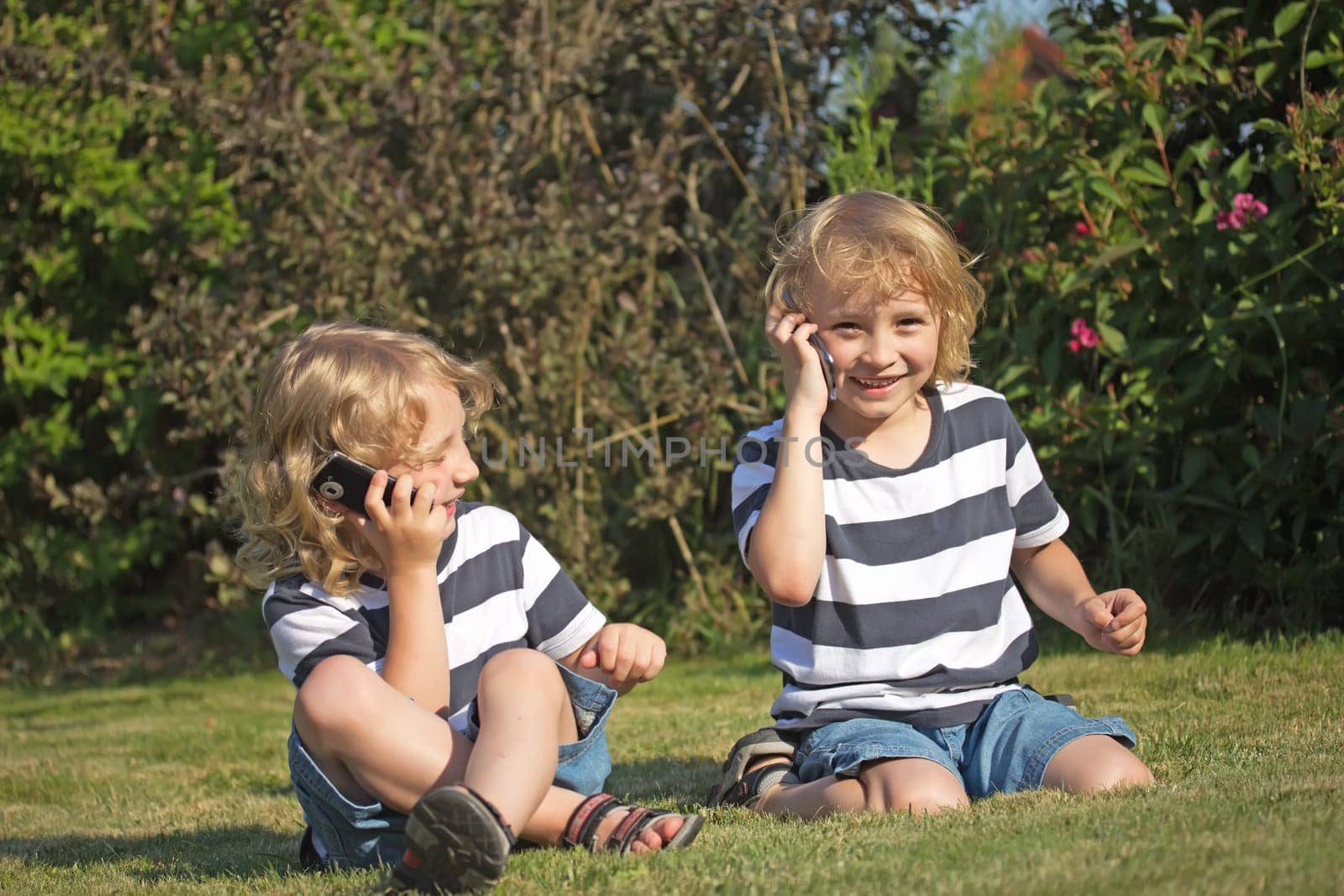 Two smiling blonde boys are sitting on the lawn and telephone.
