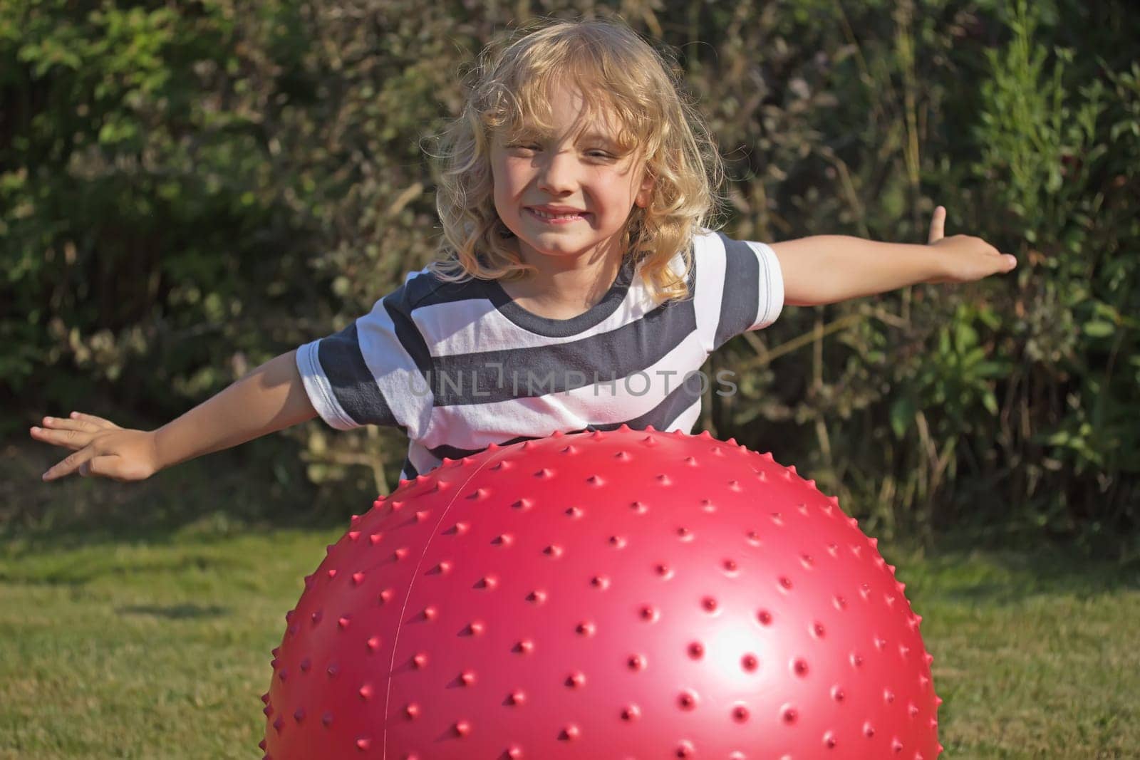 Attractive blond boy is playing with red gymnastic ball outdoors