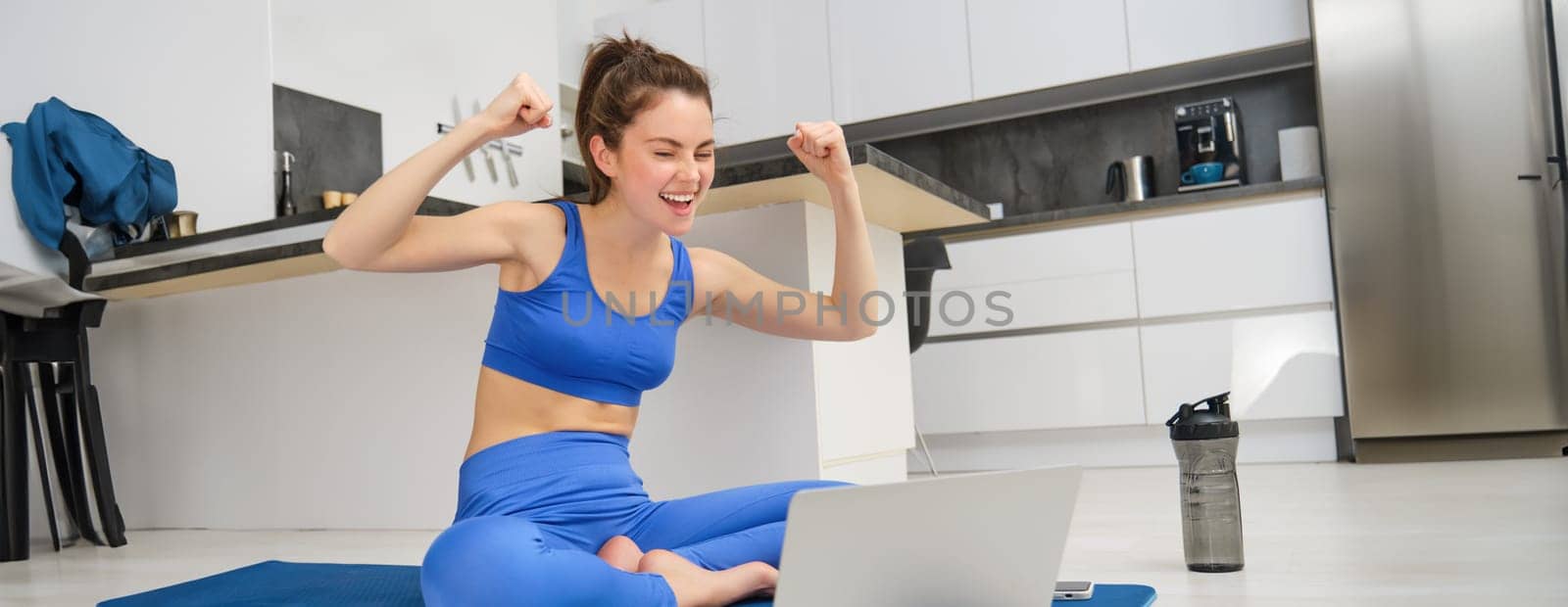 Portrait of excited young woman, sitting with laptop on mat, cheering client, giving online yoga lesson, remote fitness class, working as gym instructor from home.