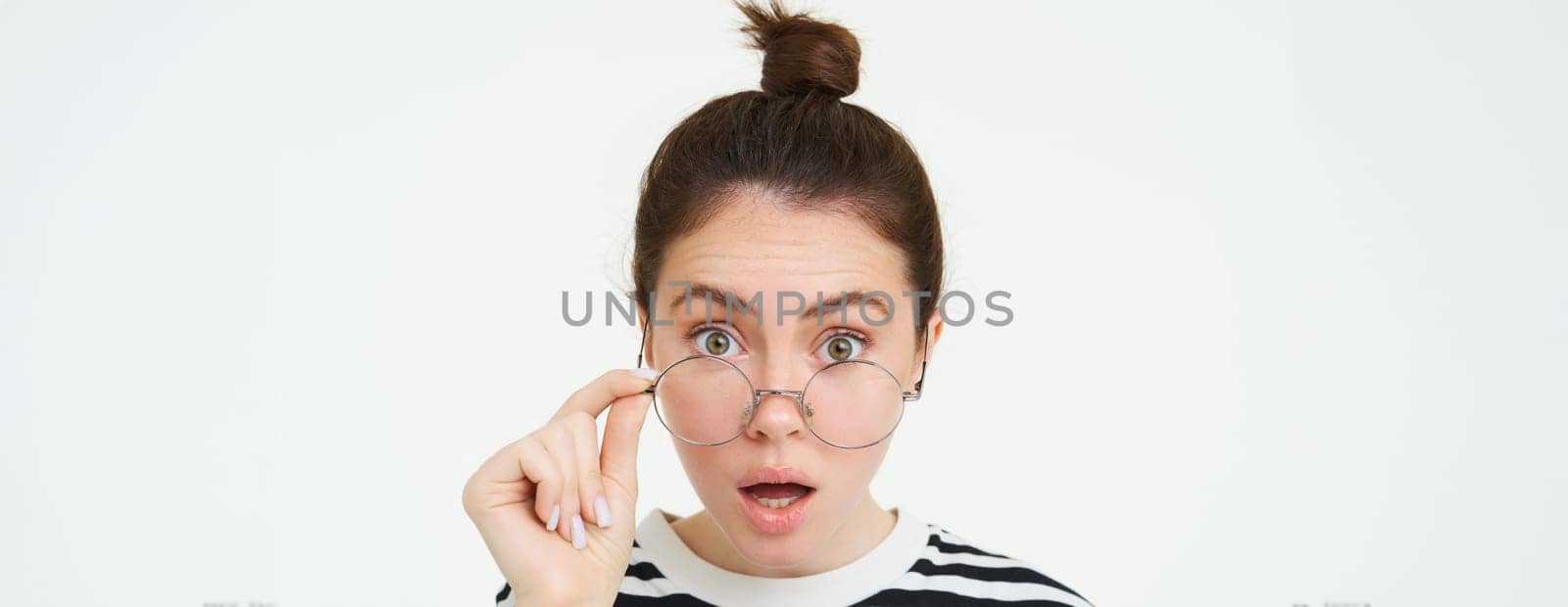 Image of woman in glasses, looks surprised, takes off eyewear, looks amazed, says wow, impressed by something, stands over white background.
