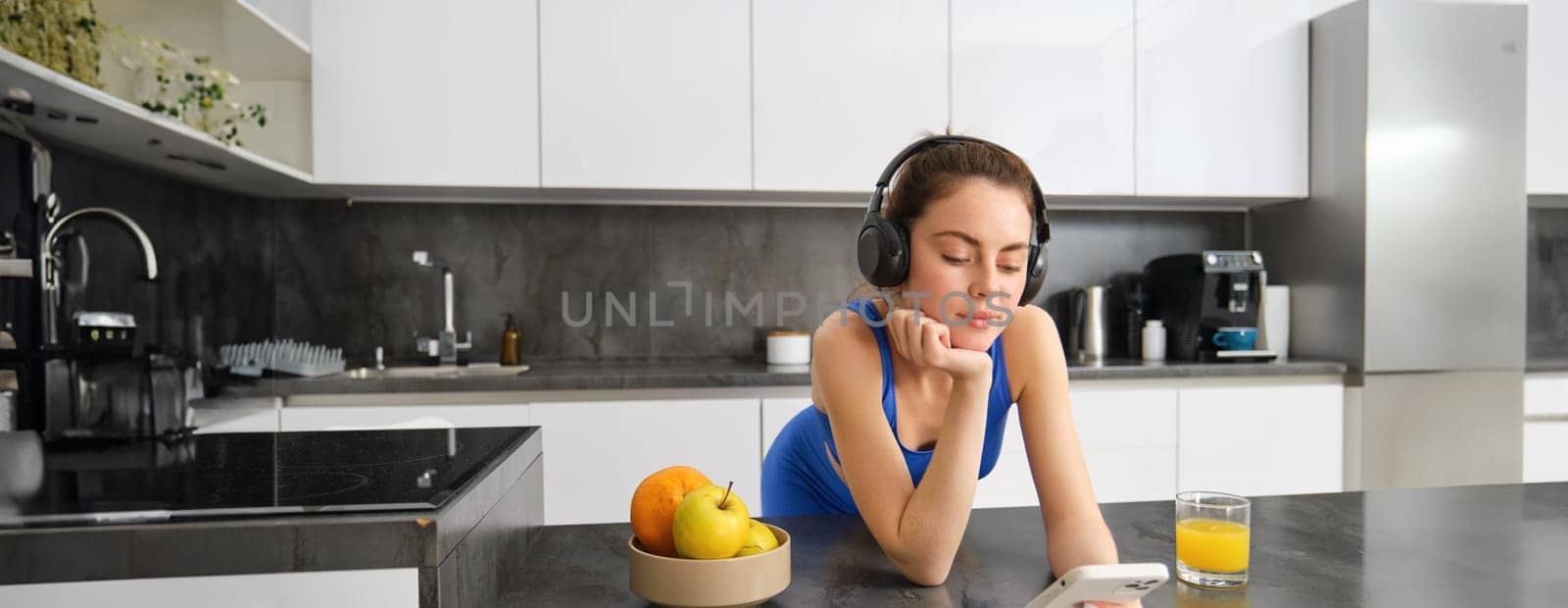 Image of happy, stylish young sports woman, standing in kitchen and drinking orange juice, listening music in headphones, using smartphone app by Benzoix