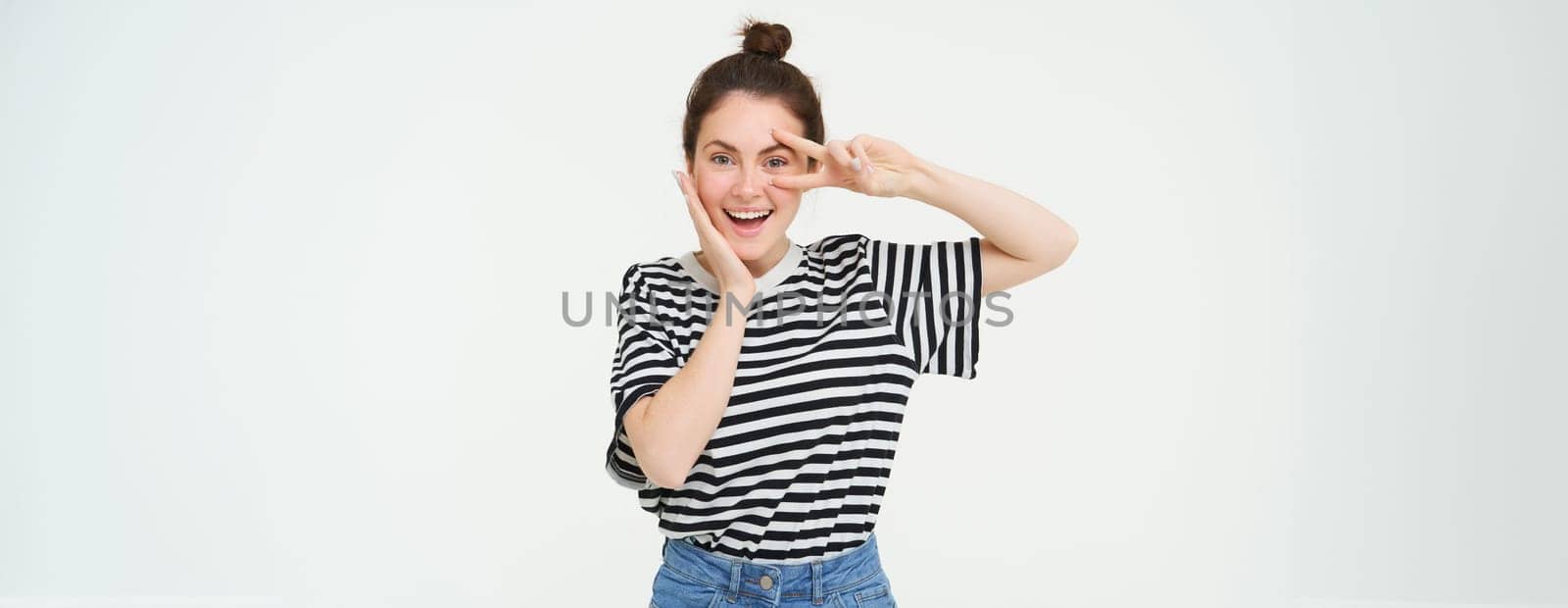 Beautiful brunette woman shows peace sign near eye and smiling, isolated against white background.