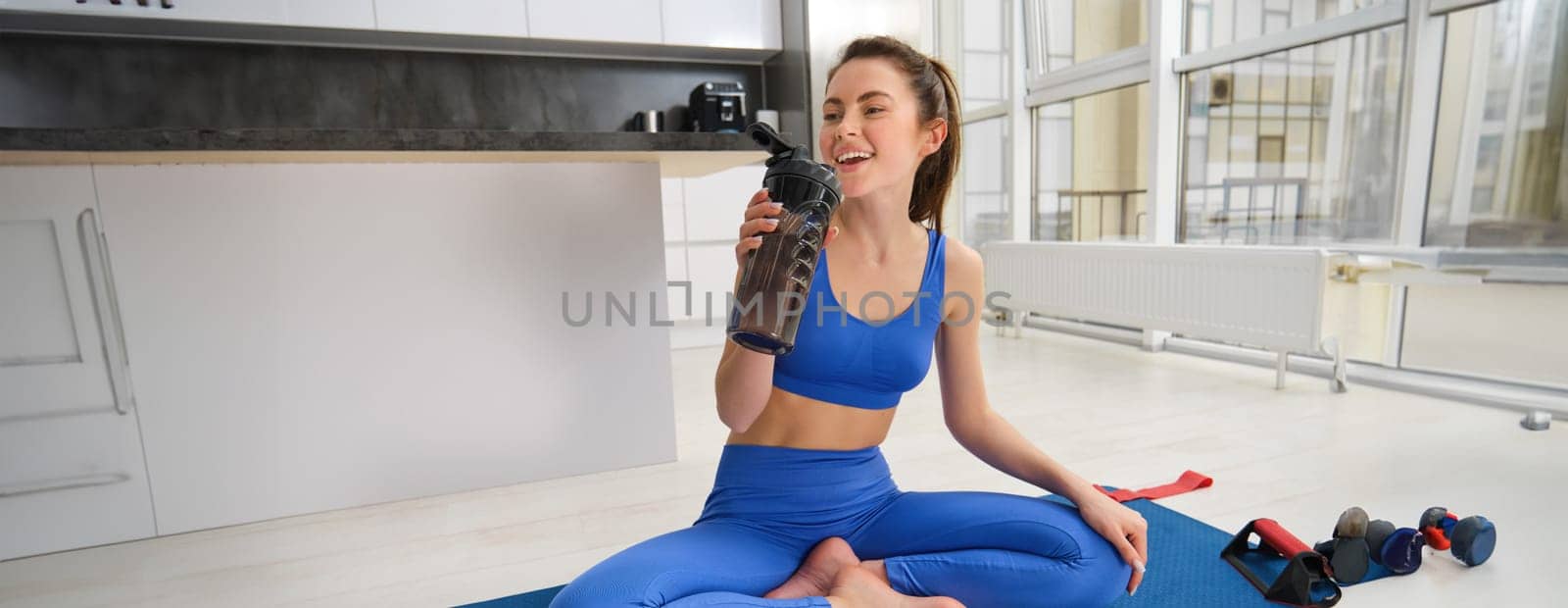 Young woman working out from home, drinking water during her sport training session in living room, sitting on yoga mat.
