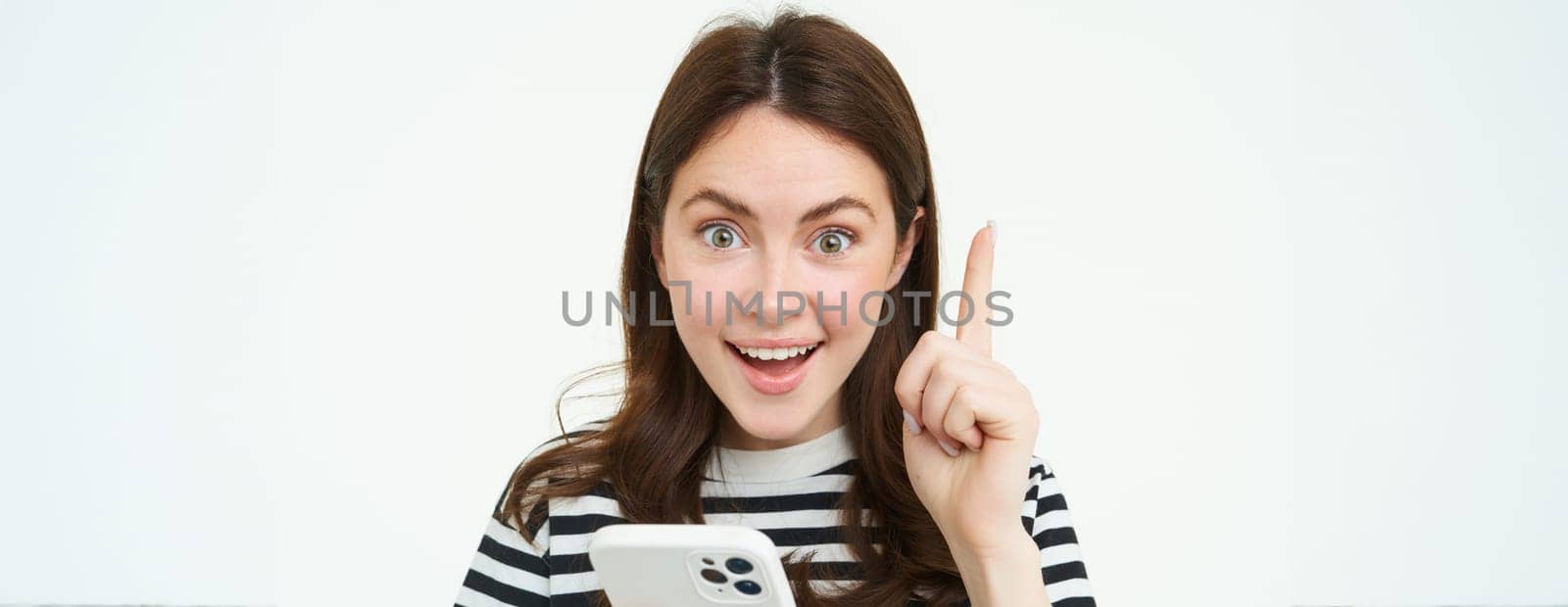 Got an idea. Smiling woman, raises her finger and holds smartphone, suggests something, makes eureka gesture, has a solution, white background by Benzoix