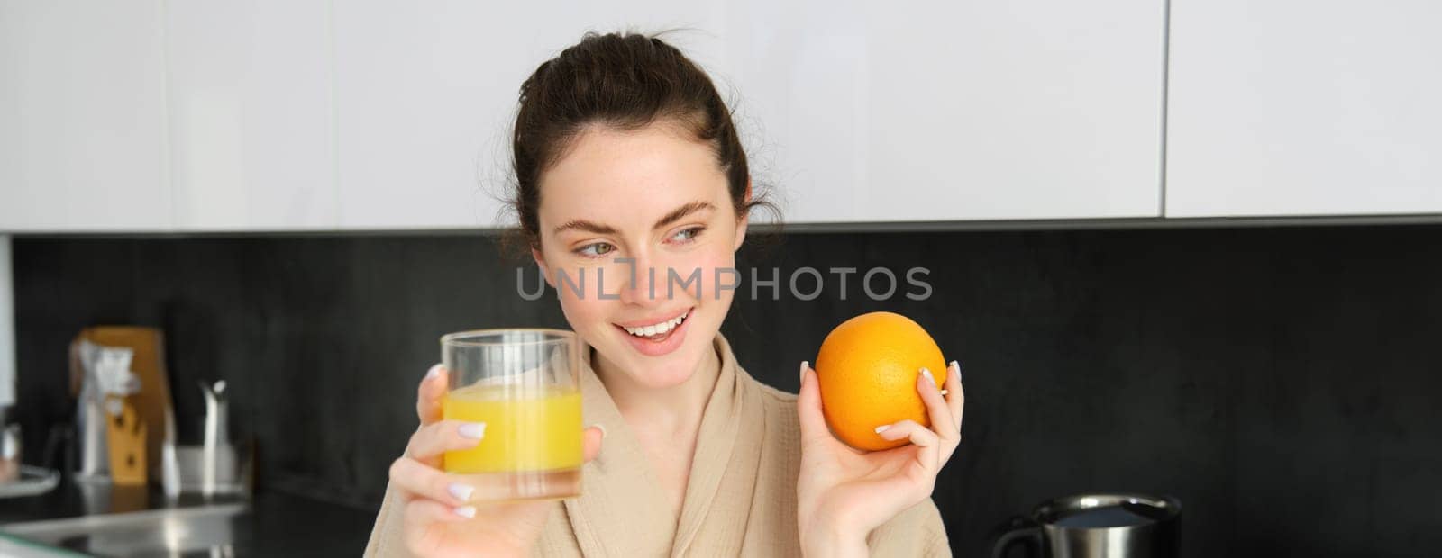 Image of good-looking healthy woman in bathrobe, drinking fresh juice, showing orange fruit, posing in kitchen by Benzoix