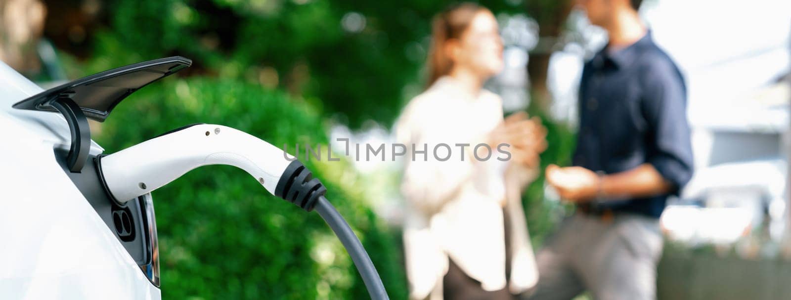 Young couple recharge electric car battery from charging station in green city park in springtime. Rechargeable EV car for sustainable environmental friendly urban travel lifestyle. Panorama Expedient