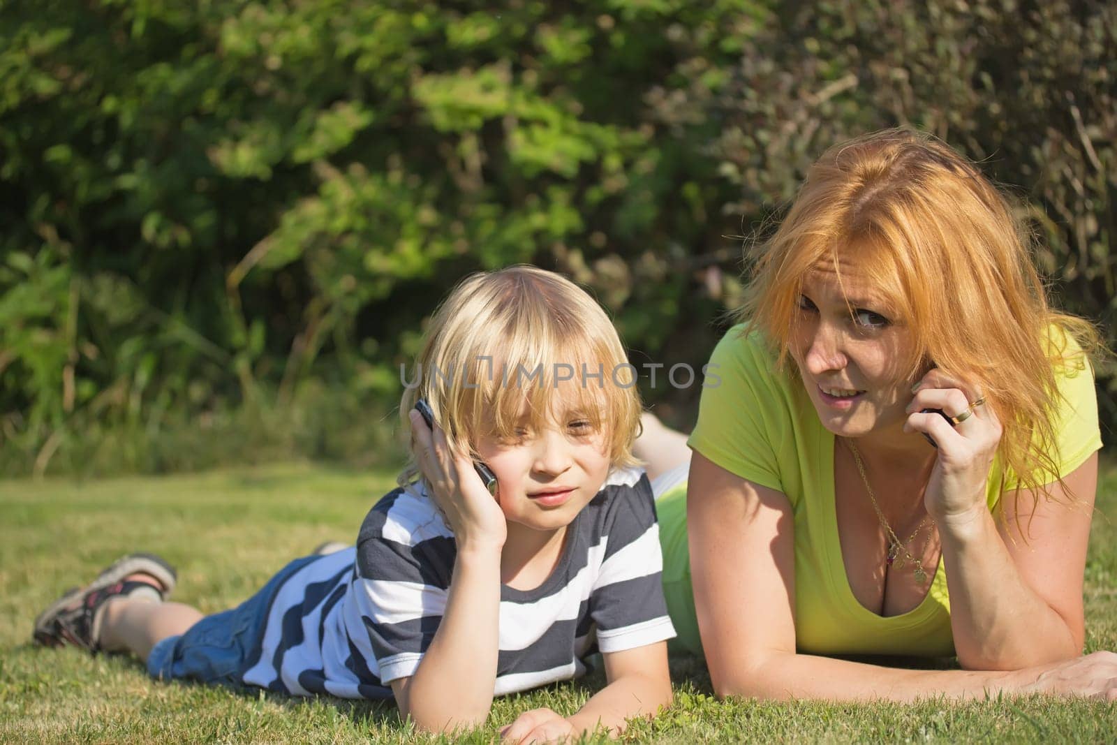 Mother with her blond hair son are lying and calling on the lawn in the garden.