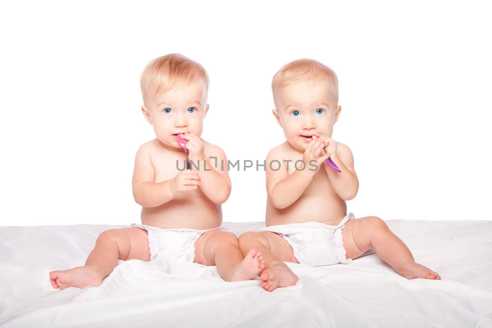 Two cute adorable infant babies with blue eyes sitting and spoons eating playing, on white.
