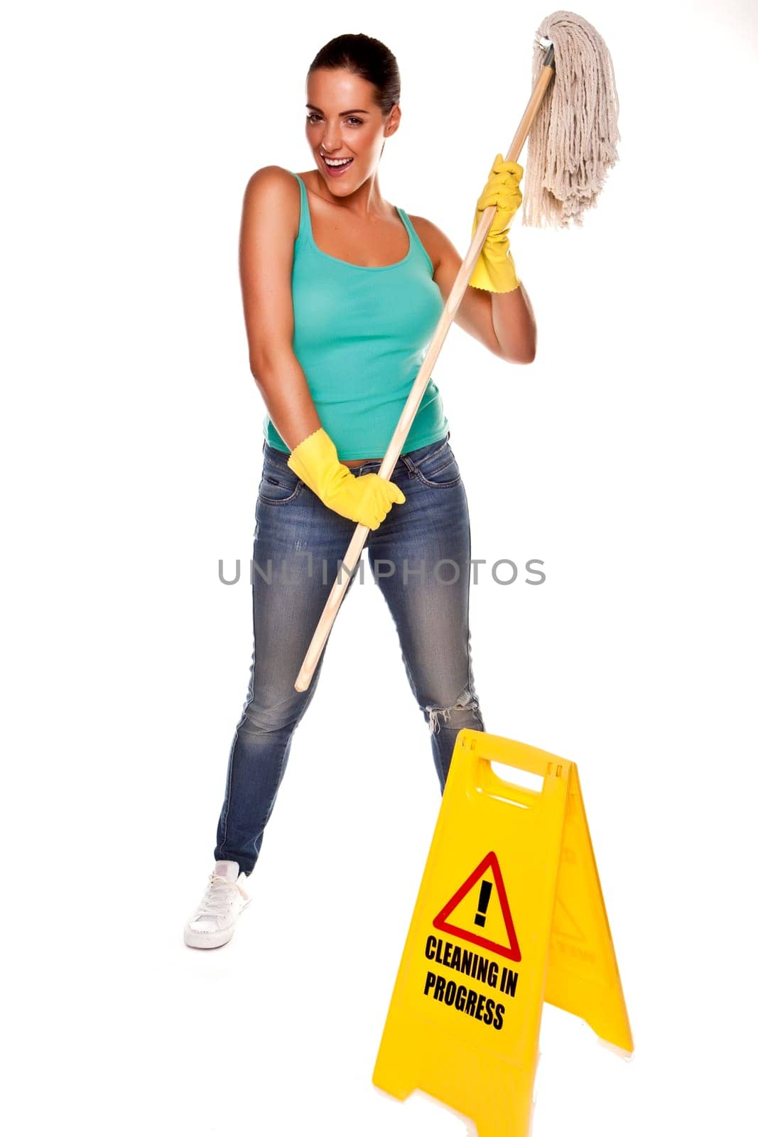 An attractive young woman with brunet hair wearing a blue vest top and yellow rubber cleaning gloves and doing some cleaning on a white background .