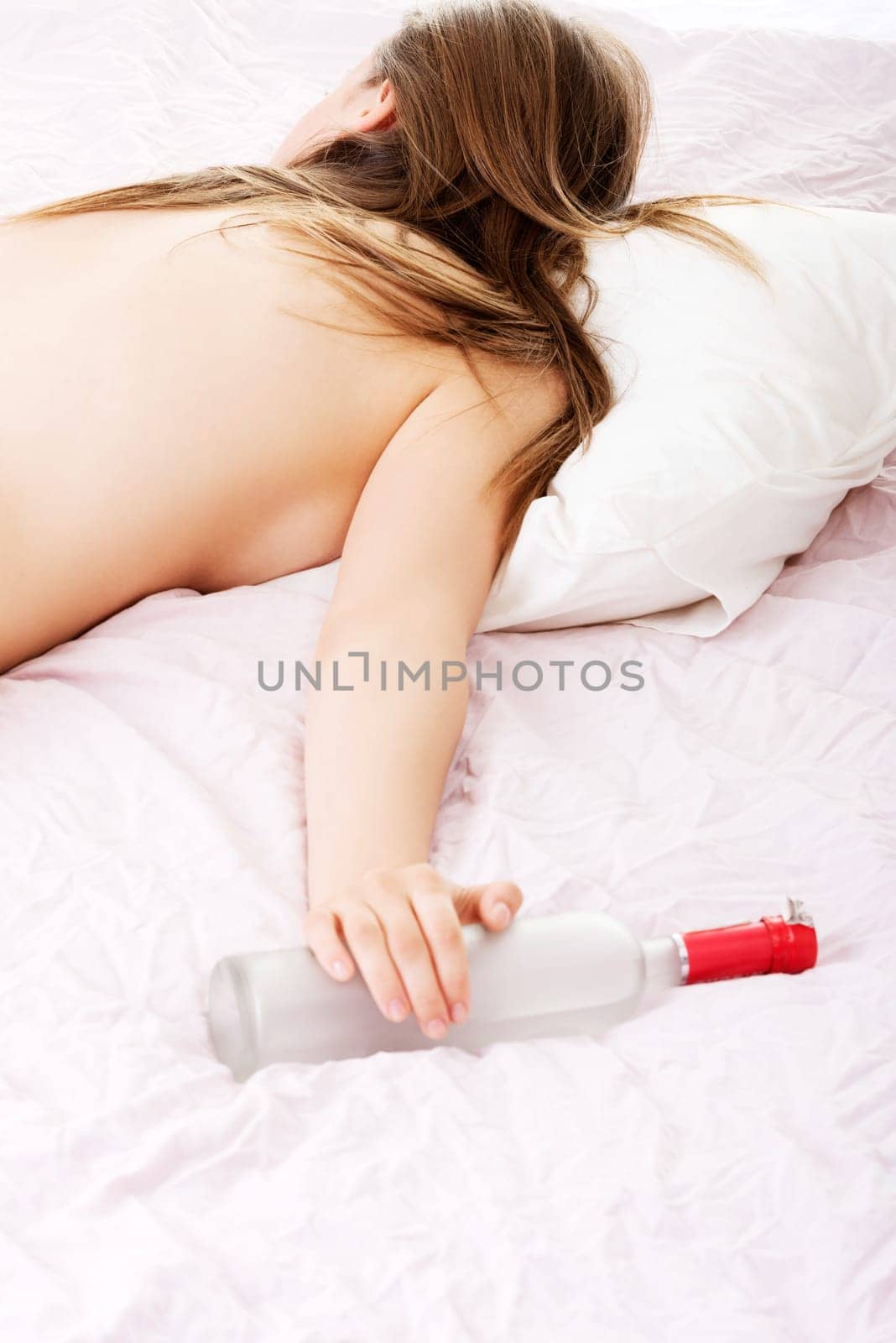 Drunk young topless woman sleeping on bed with bottle of vodka in hand. Isolated on white