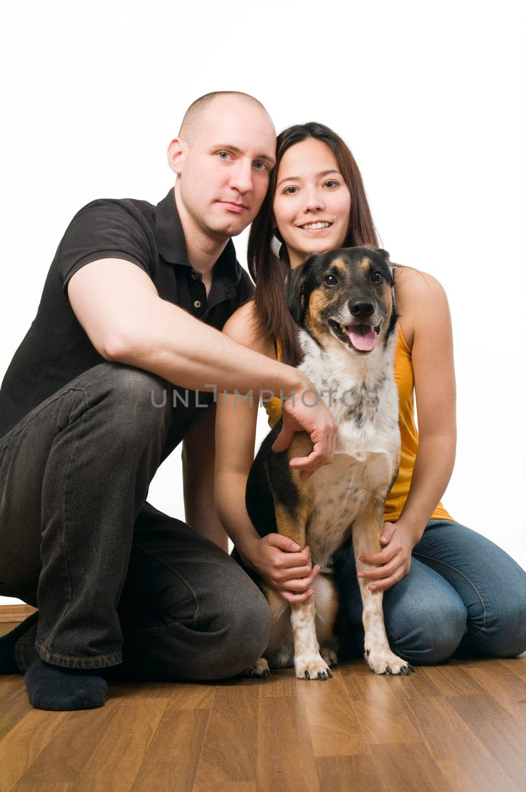 Full body portrait of a young couple with dog sitting on wooden floor in front of a white background