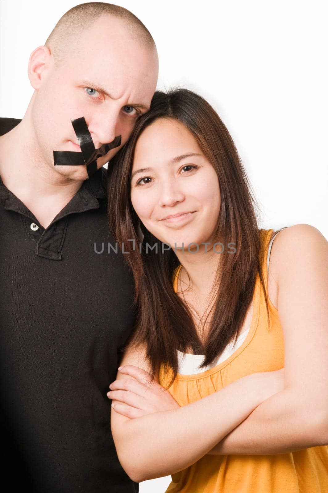 Upper body portrait of a young couple against a white background  Man has mouth glued crosswise with duct tape  Woman looks mischievously smiling with crossed arms into the camera