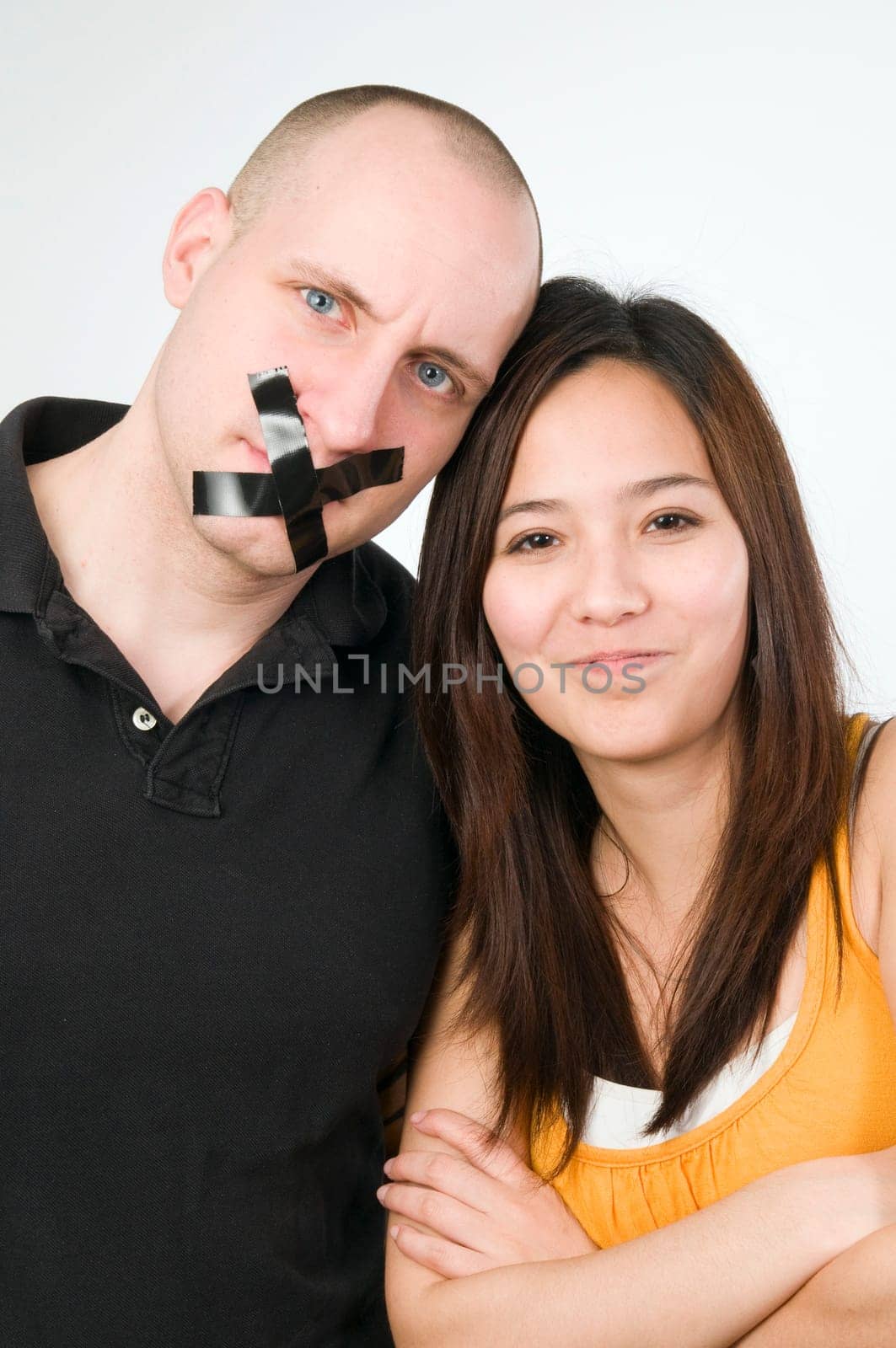 Upper body portrait of a young couple against a white background  Man has mouth glued crosswise with duct tape  Woman looks mischievously smiling with crossed arms into the camera