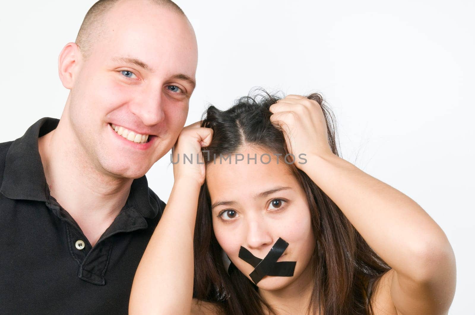 Head-and-shoulder portrait of a young couple against a white background  Woman has her mouth glued crosswise and looks questioningly and hair ruffling into the camera  Man looks into the camera with a broad grin