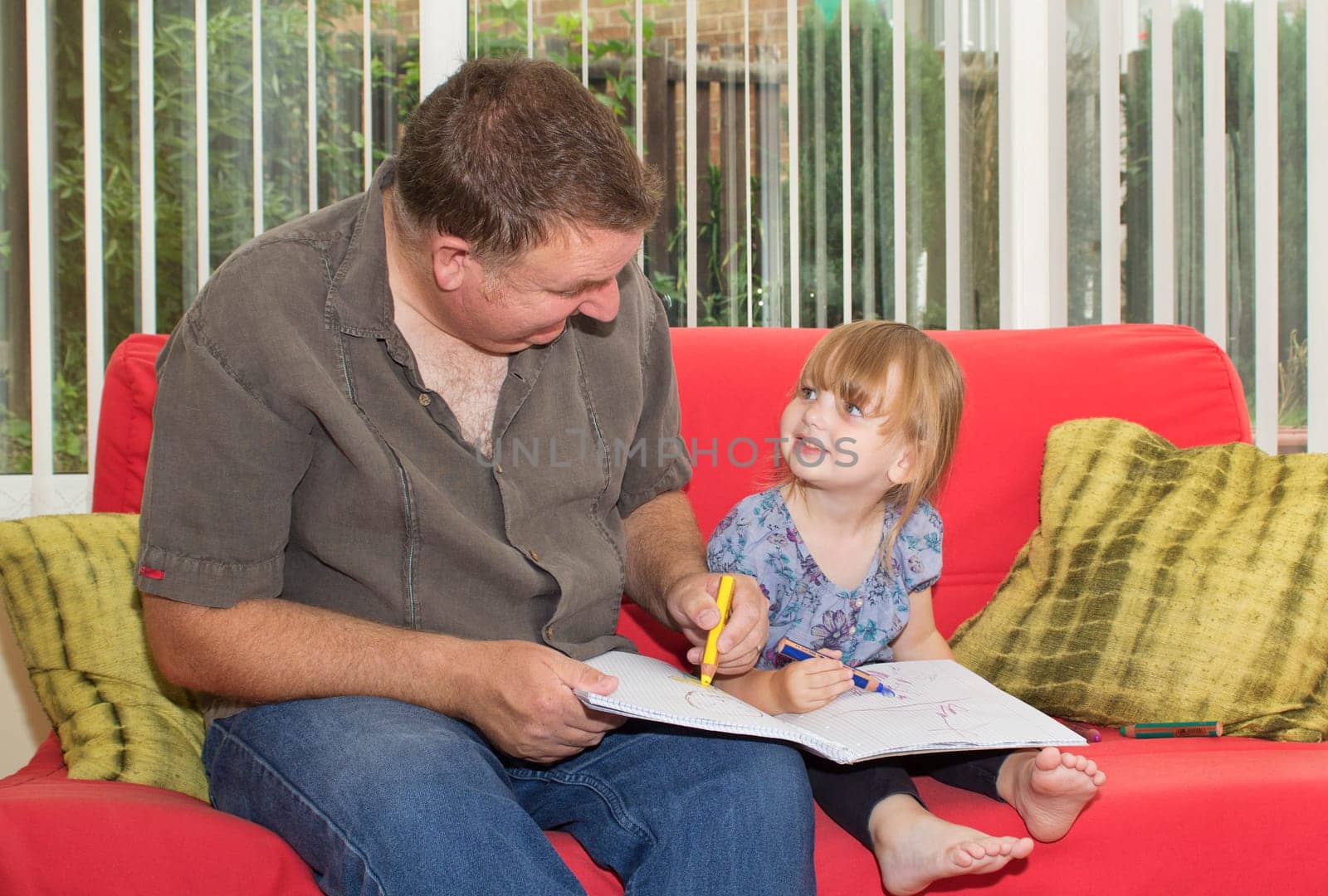 Father and daughter sitting on a sofa, drawing a picture together. The little girl is smiling up at her dad.