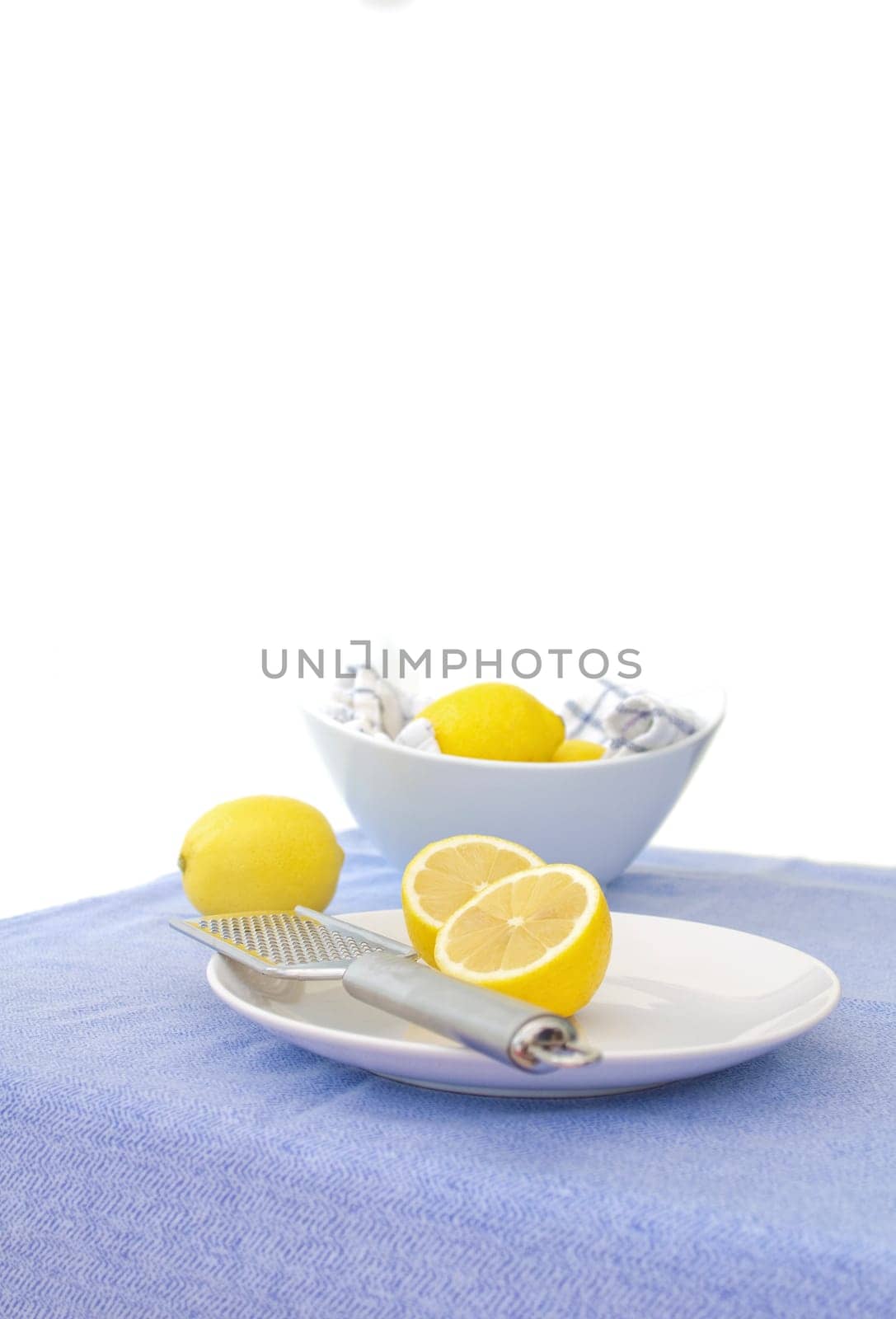 cut lemons on a dish with a lemon zester, with a bowl of lemons in the background. The tablecloth is a light blue, to compliment the yellow of the lemons.