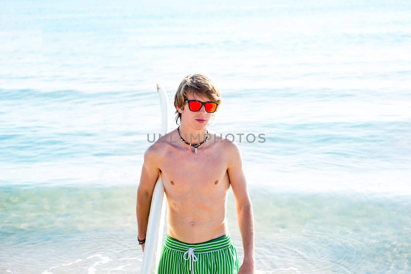 Boy teenager with surfboard on beach shore and orange sunglasses