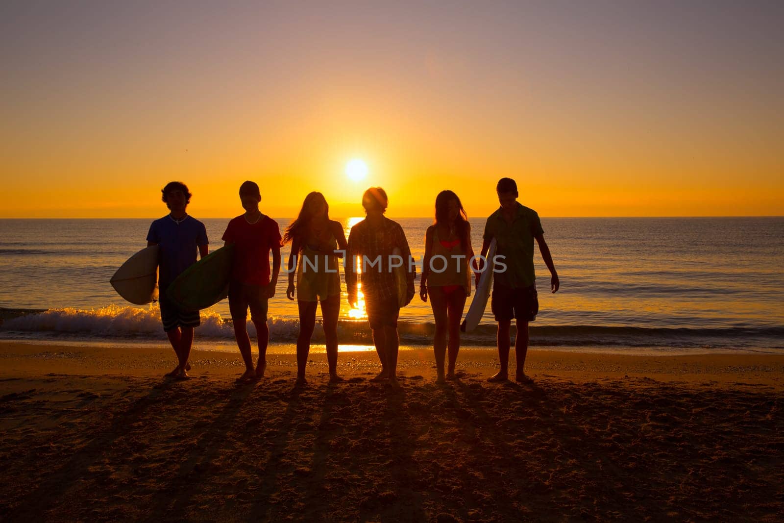 Surfers teen boys and girls group walking on beach at sunshine sunset backlight