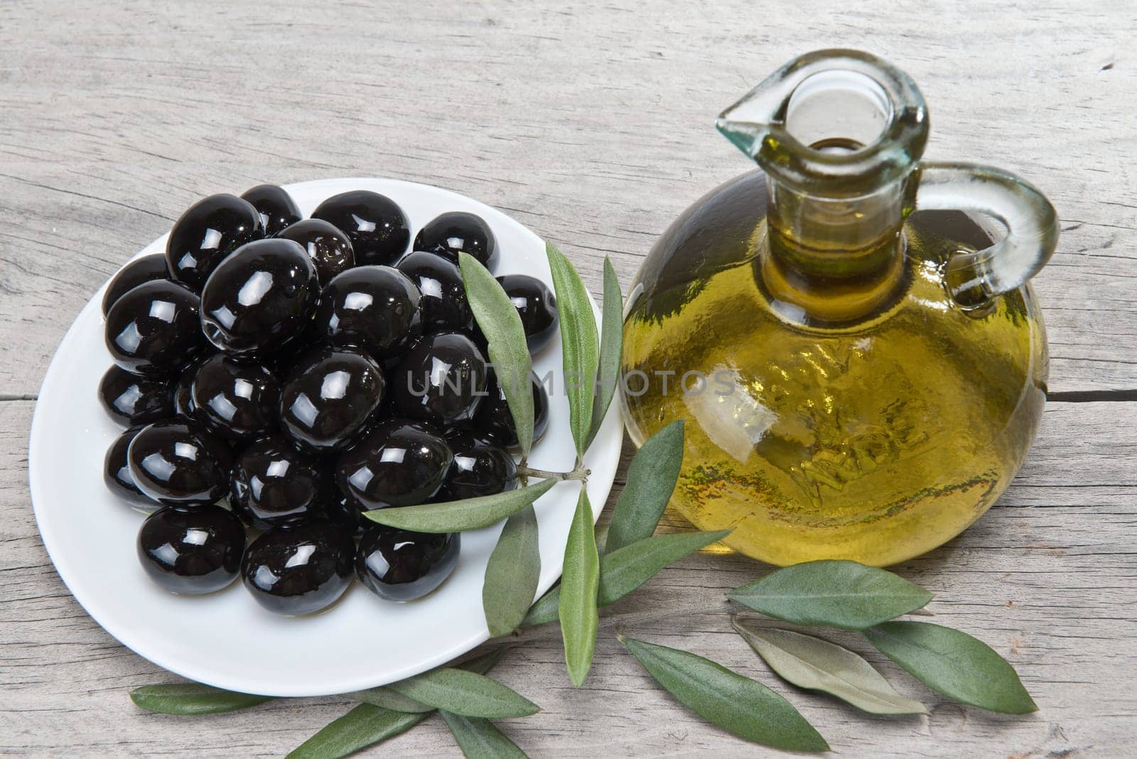 A jar with olive oil ans a plate with black olives on a wooden surface.