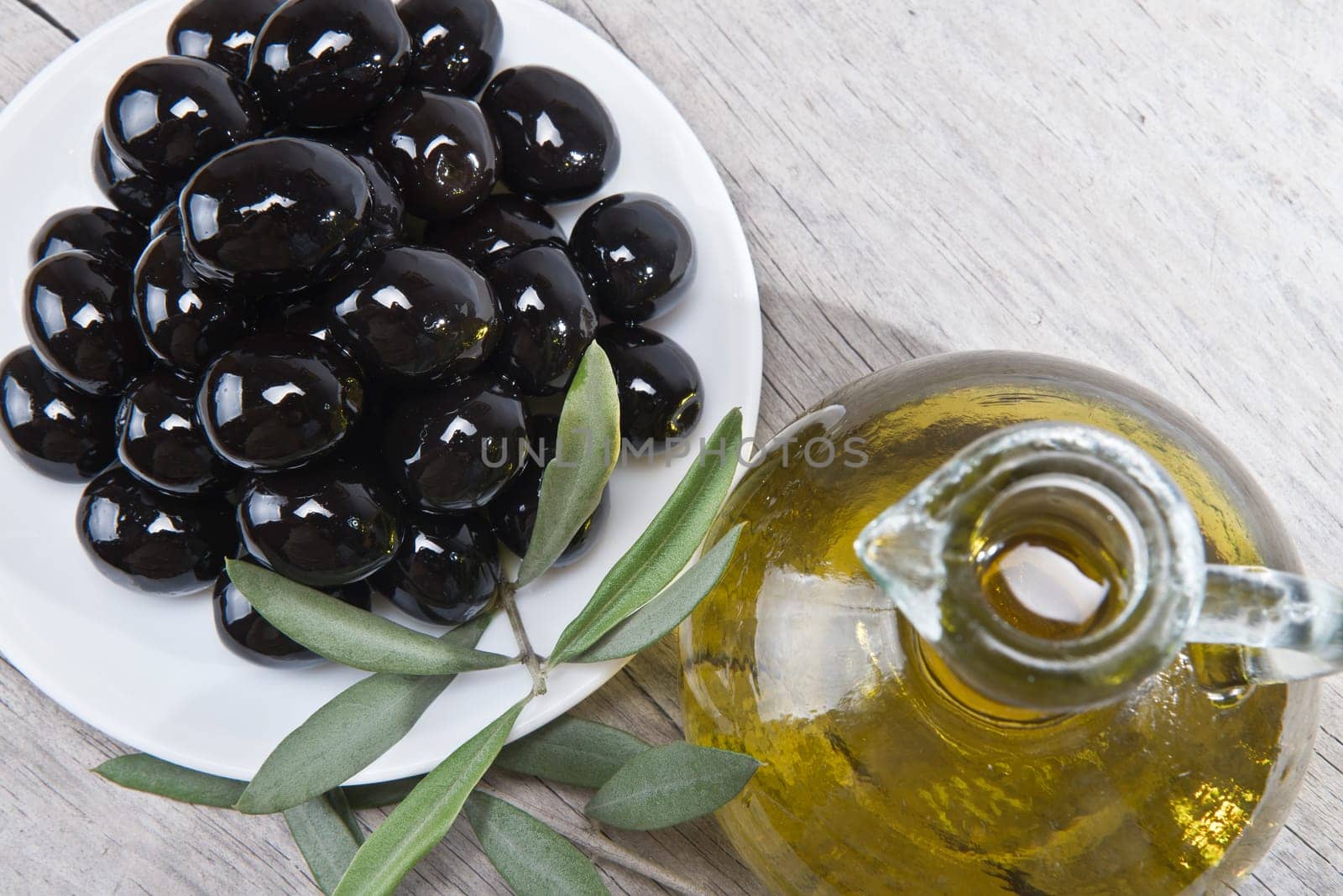 A jar with olive oil ans a plate with black olives on a wooden surface.