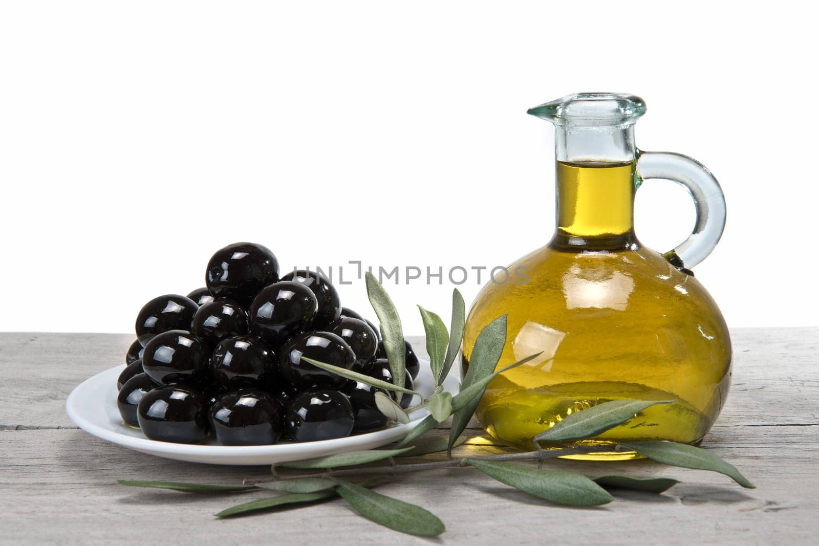 A jar with olive oil ans a plate with black olives on a wooden surface.