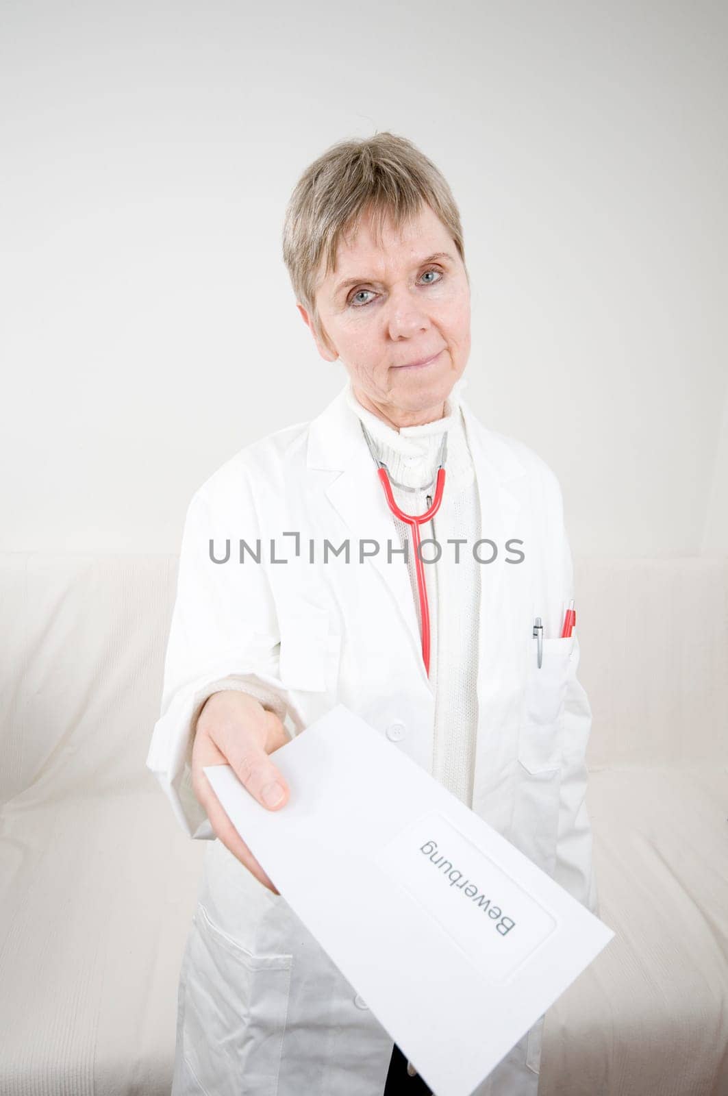 elderly woman in white doctor's coat and red stethoscope around her neck hands over a letter wrap with application inscription towards the camera