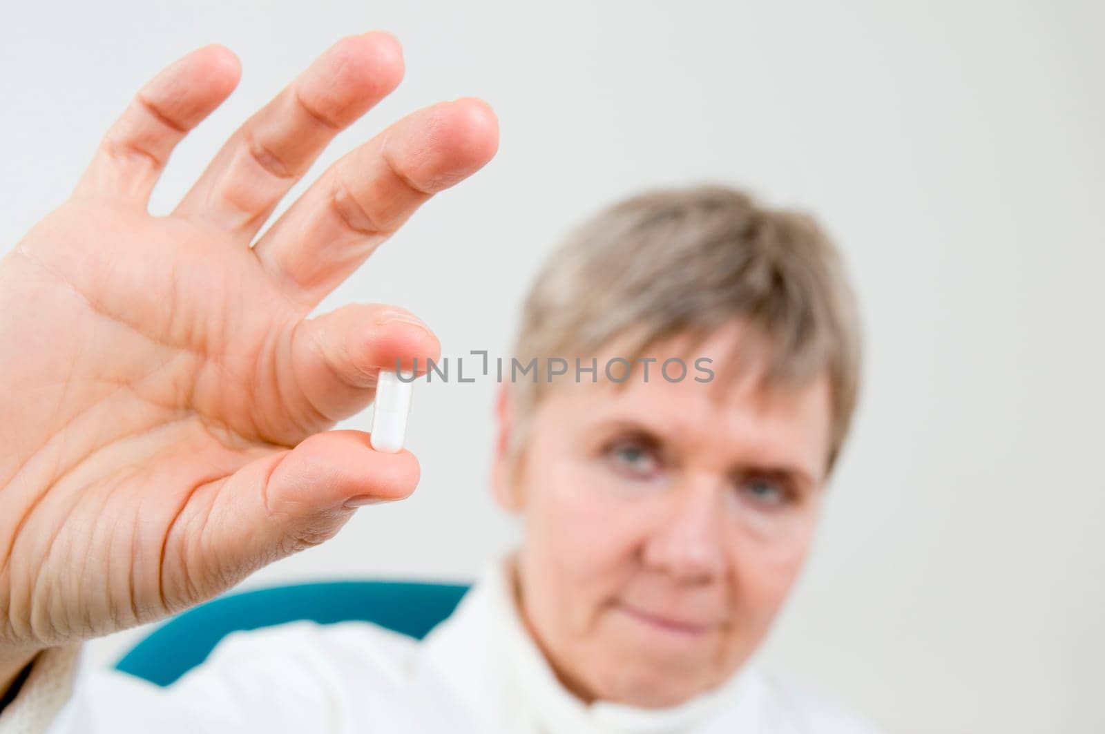 Head-and-shoulder shot of an elderly woman in a white coat holds a white medicine capsule between two fingers and looks into the camera