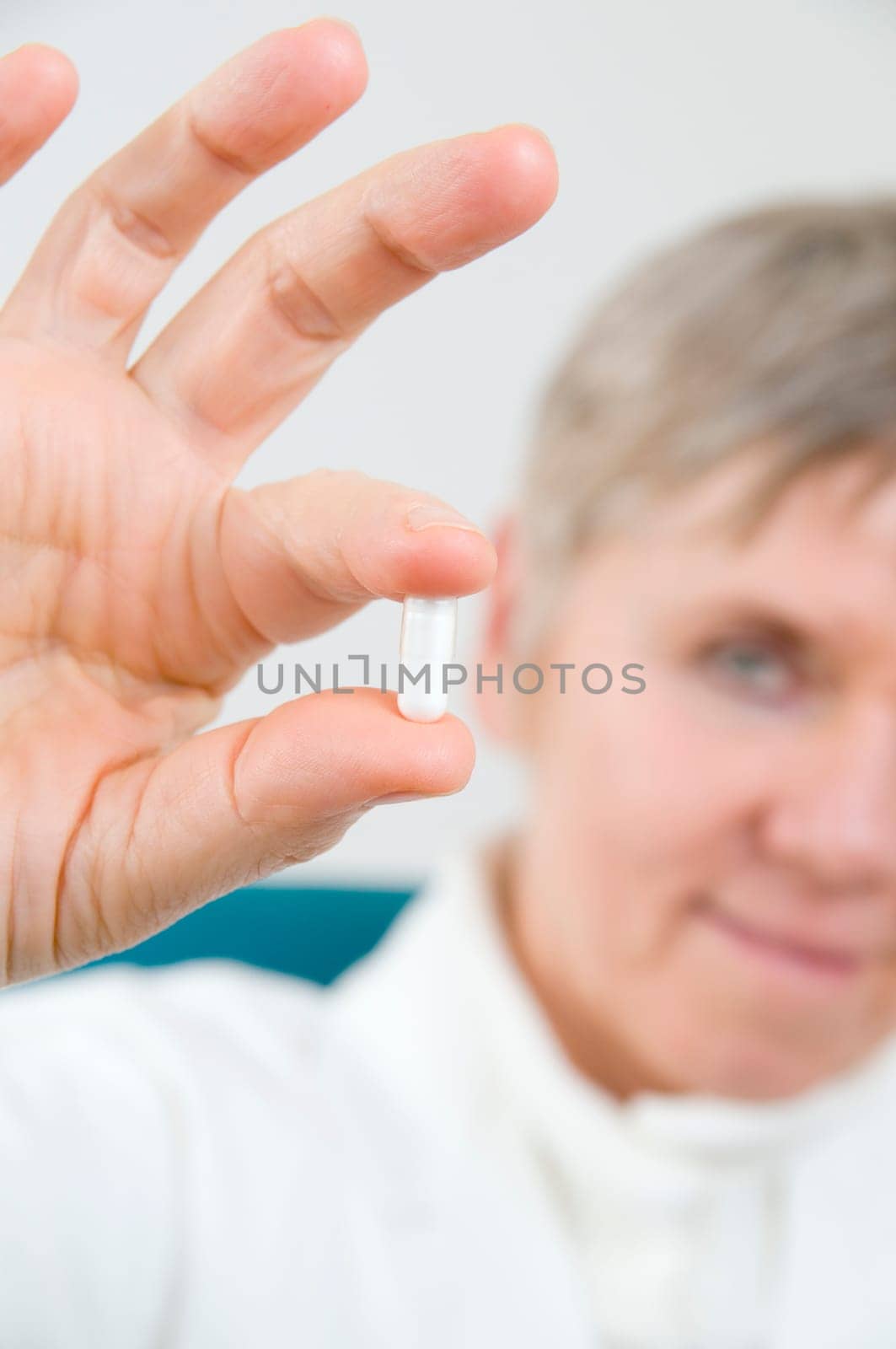 Head-and-shoulder shot of an elderly woman in a white coat holds a white medicine capsule between two fingers and looks into the camera